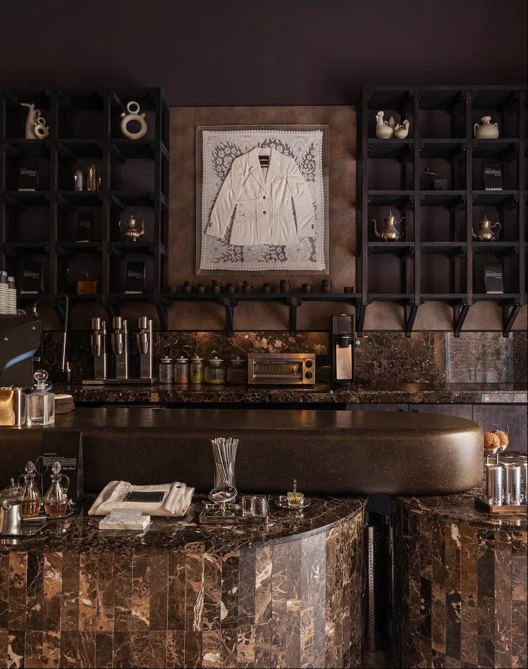 Barra de mármol de la cafetería de Casona, con estanterías oscuras, cafeteras de plata y una chaqueta blanca enmarcada