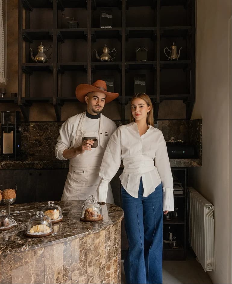 Barista con sombrero y delantal junto a Carmela, fundadora de Casona, detrás de la barra de mármol con vitrinas de bakery