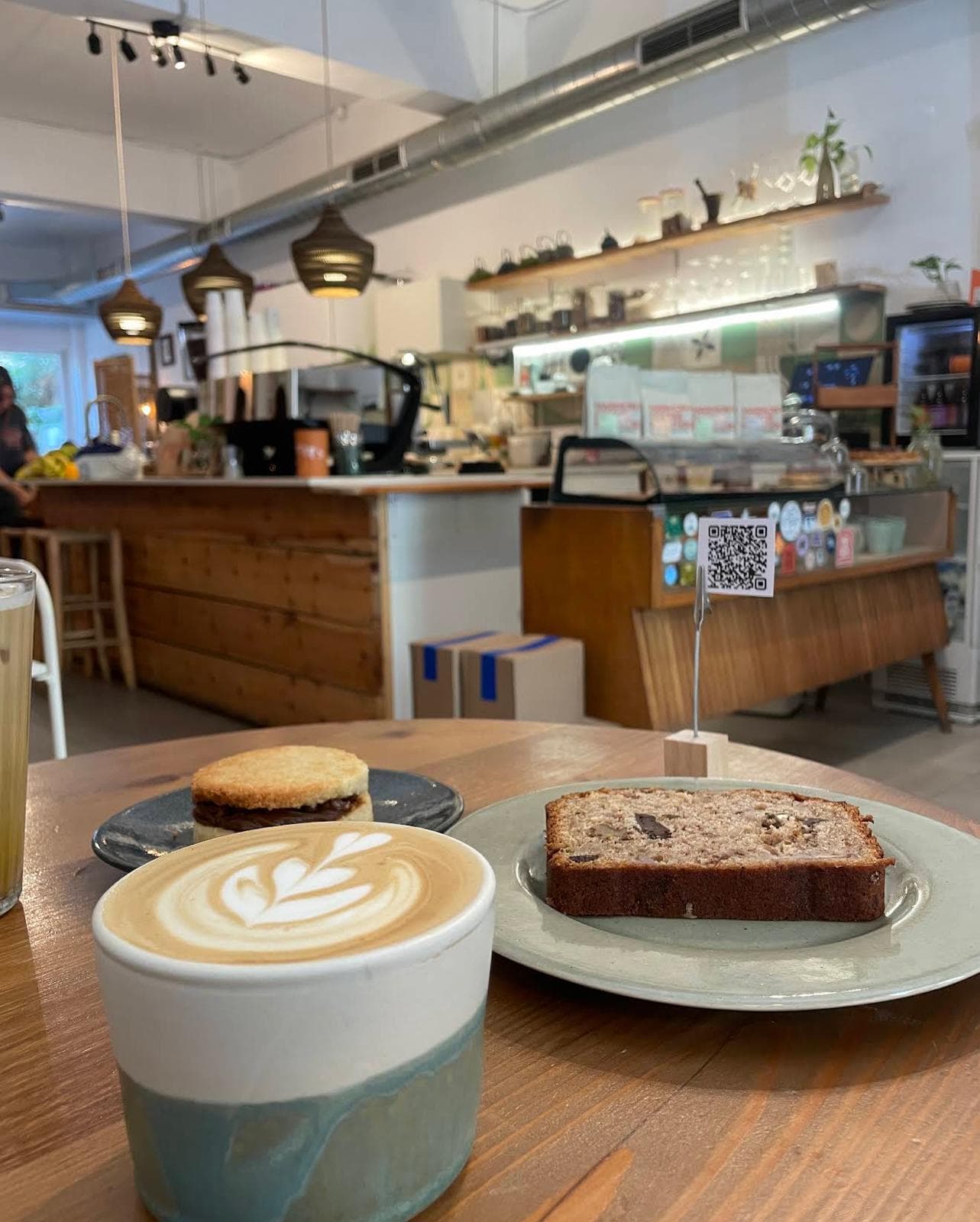 Flat white con latte art, alfajor y banana bread sobre una mesa de madera en Orígenes, con la barra al fondo